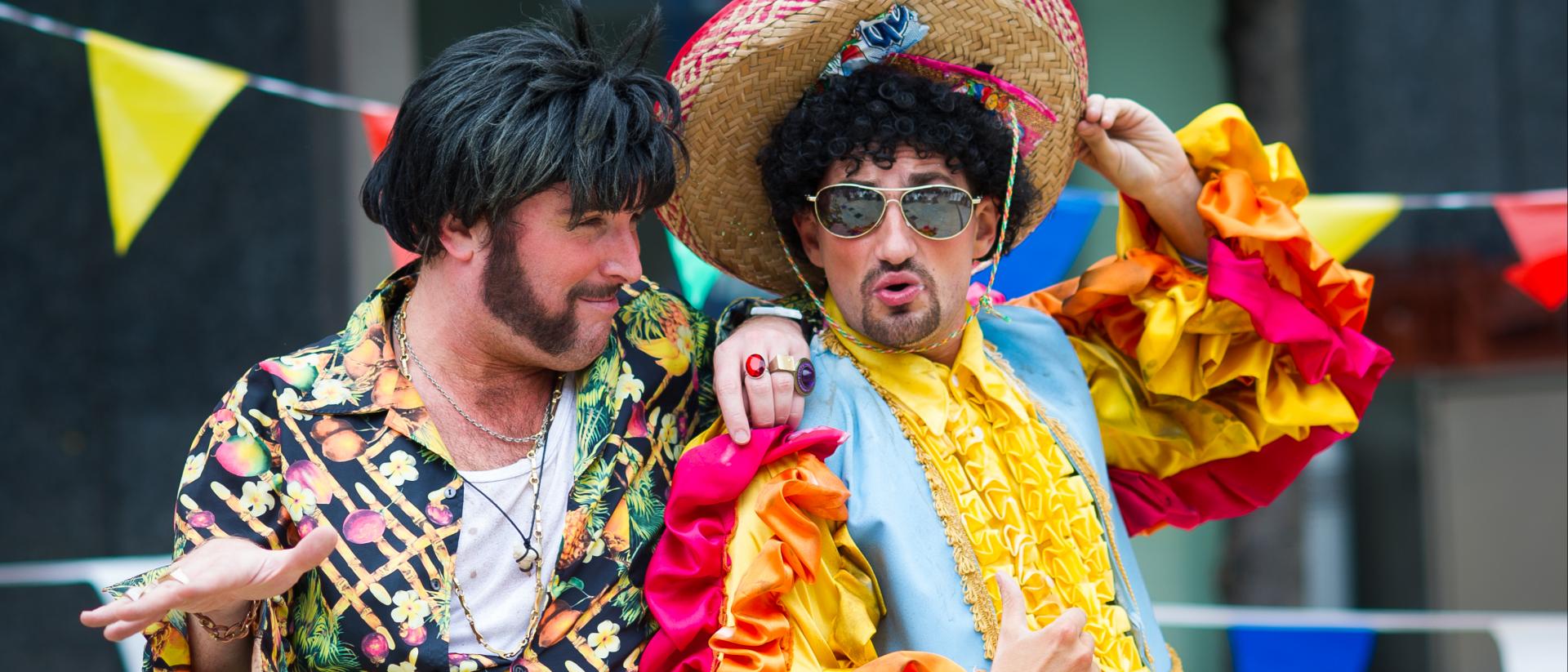 A man with black quiff, huge sideburns and wearing a loud 70s shirt is talking to another man wearing a colourful Mariachi costume, aviator specs and sombrero.
