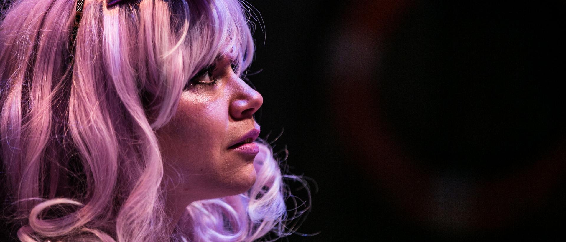 A girl with wavy purple hair and a purple bow looks wistfully out into the audience. 