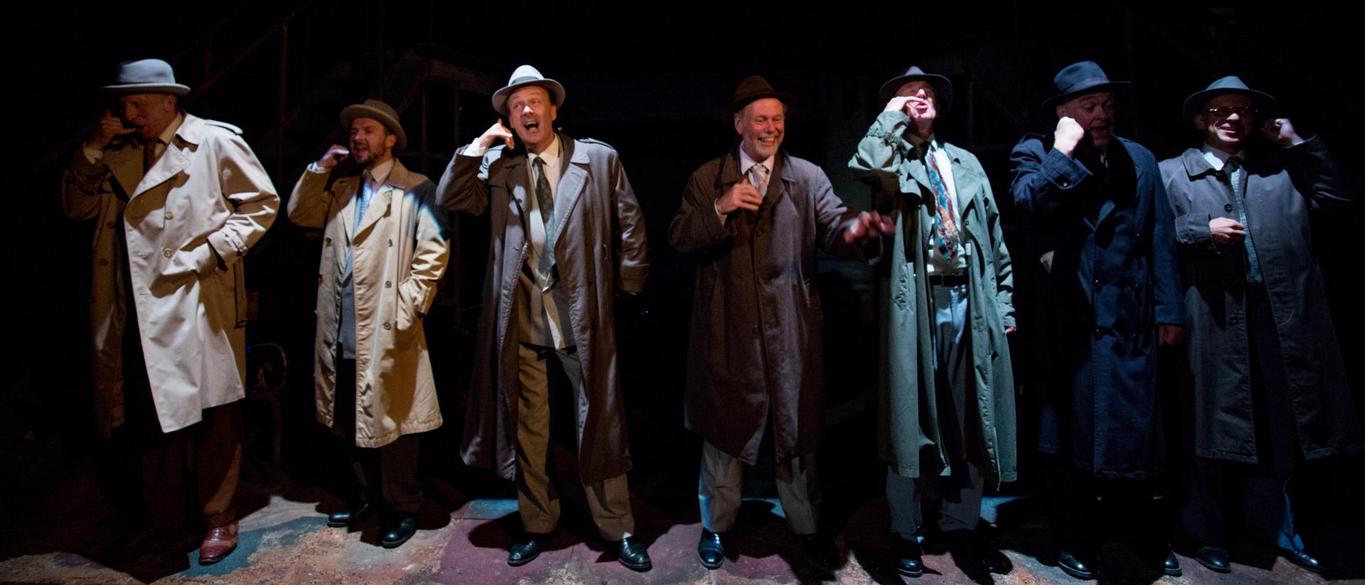 Seven men in coats and hats stand in a line and mime making a phone call. They are atmospherically lit.