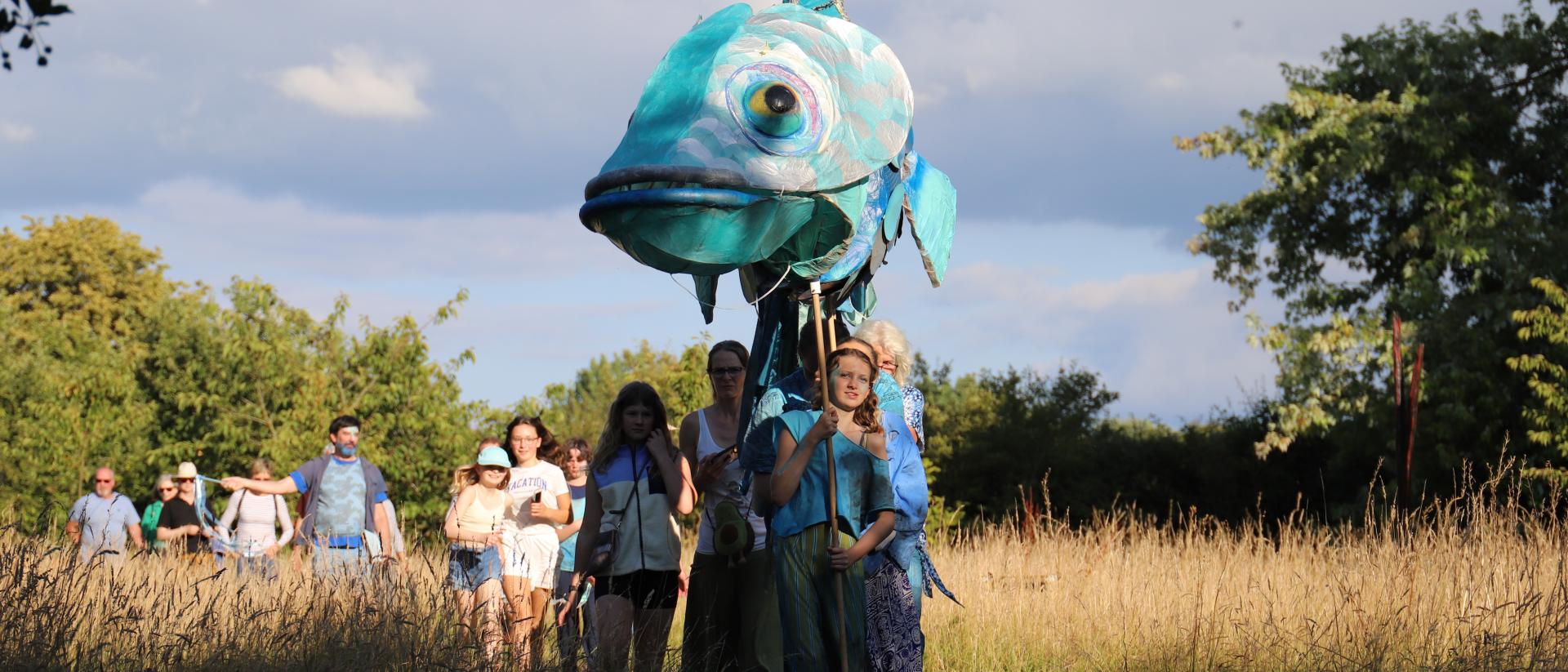 Large fish puppet parades through a meadow