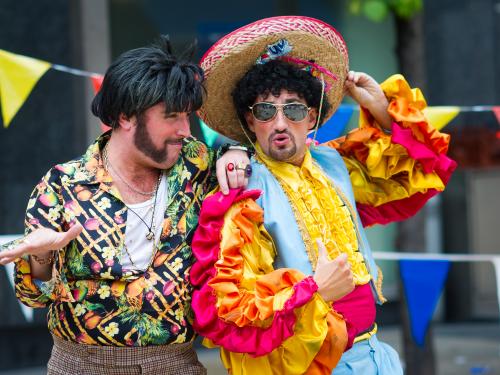 A man with black quiff, huge sideburns and wearing a loud 70s shirt is talking to another man wearing a colourful Mariachi costume, aviator specs and sombrero.
