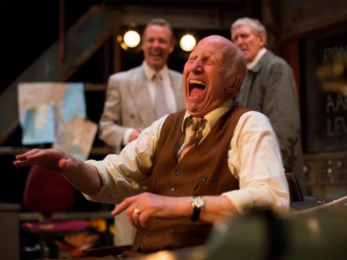 A man wearing a brown 70s waist coat, beige shirt and patterned tie is laughing in the foreground. Two other men wearing grey are seen behind, looking at him.