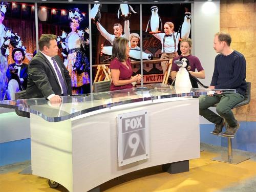 Two members of the cast - a man in a blue jumper and grey trousers and a woman wearing a purple top holding a penguin puppet - sit in the Fox 9 TV studio and are being interviewed by two presenters.