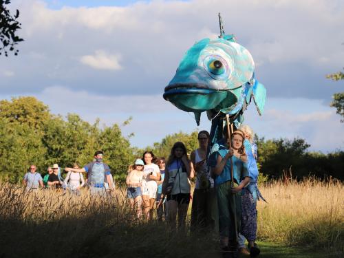 Large fish puppet parades through a meadow