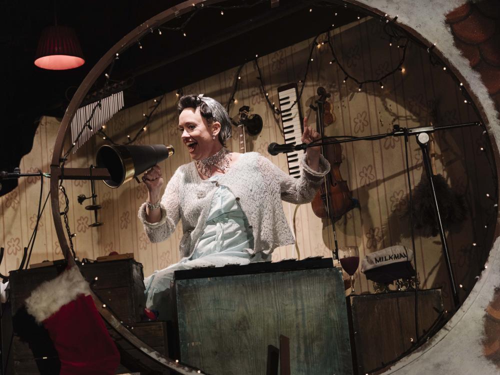 A lady is seen through a window, surrounded by instruments. She looks happy and is using a loud haler to speak.