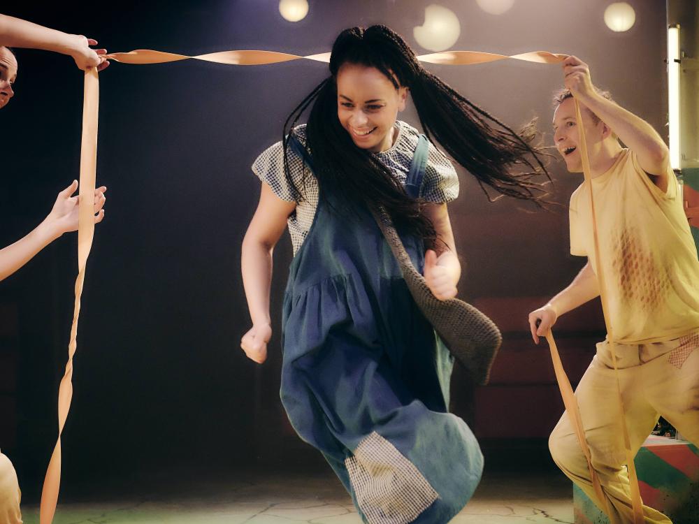 A smiling woman runs through a yellow door made of elastic and held up by two other smiley humans.