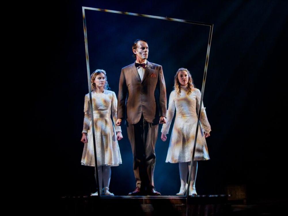 A man in a brown suit stands behind a window frame in low light. Two ghostly women dressed in white stand behind him.