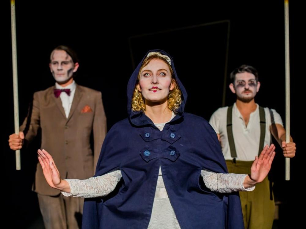 A woman with pale eyes steps through a door into an apartment block. Two men create the outline door with long poles.