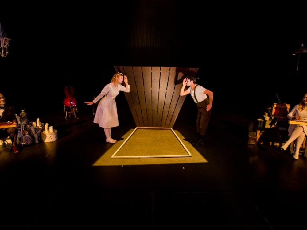 A woman and a man communicate through the spy hole in a door. Musicians play glockenspiels at the side of the stage.