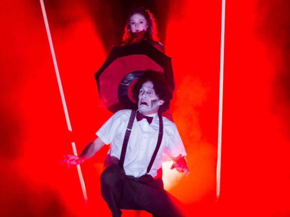 A man screams, bathed in red light. A woman spins a red and black umbrella behind him.